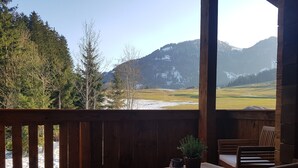 Een houten terras met een tafel en stoelen, met uitzicht op een bergachtig landschap met sneeuwvlekken en bomen.