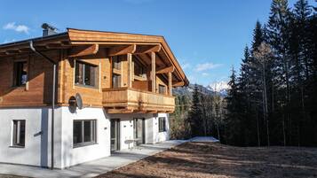 Une maison moderne en bois, dotée d’un balcon, de grandes fenêtres et offrant une vue sur une forêt et des montagnes.