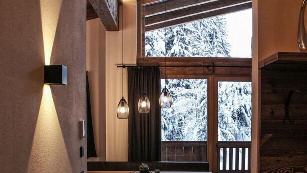 Een gezellige eethoek met een houten tafel en stoelen, een moderne hanglamp en zicht op besneeuwde bomen door grote ramen.