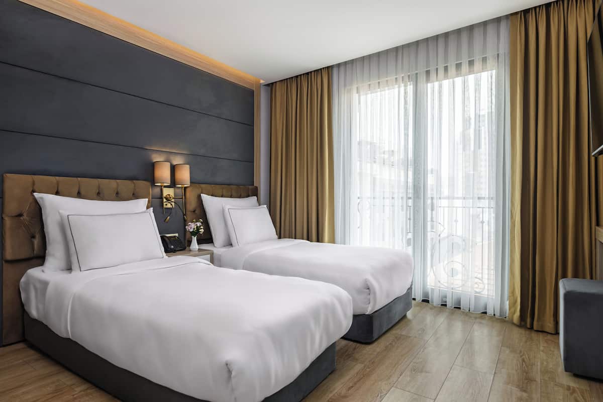 Habitación de hotel con dos camas, una mesita de noche con lámparas, un ventanal con cortinas y un balcón.