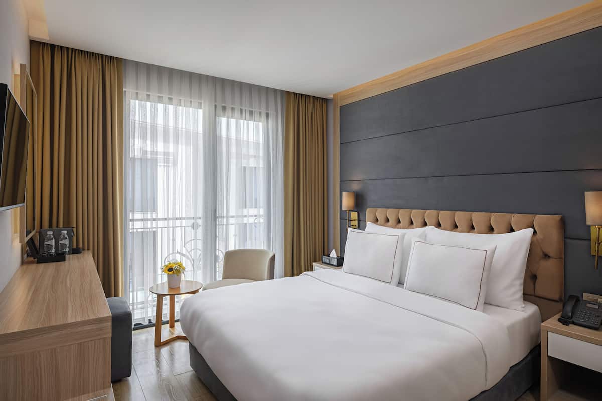 Habitación de hotel con una cama grande, una mesita de noche, una lámpara, una silla, una pequeña mesa con un jarrón de flores y un televisor a la izquierda.
