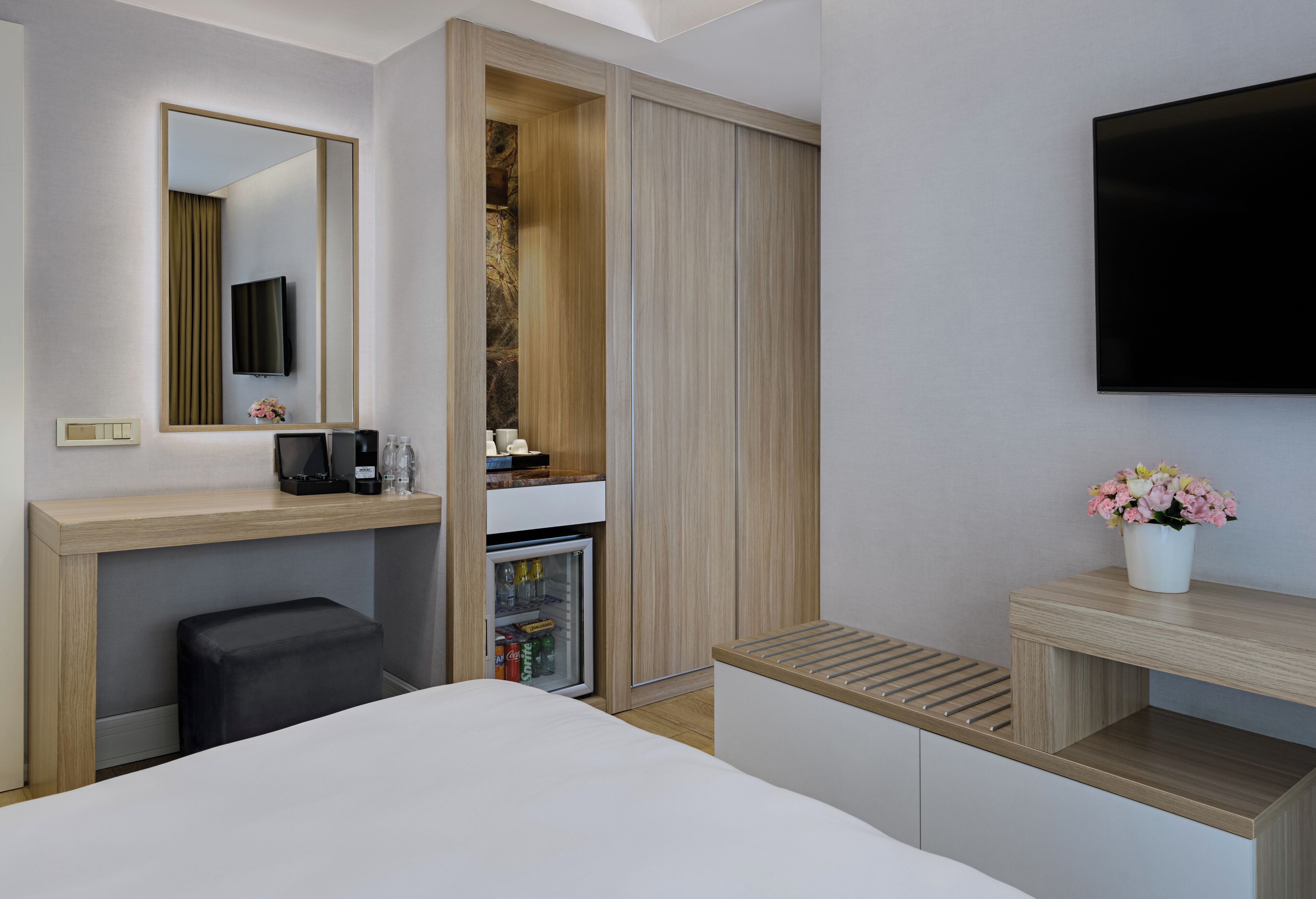 A modern hotel room with a bed, a TV mounted on the wall, a minibar, a small seating area, and a mirror.