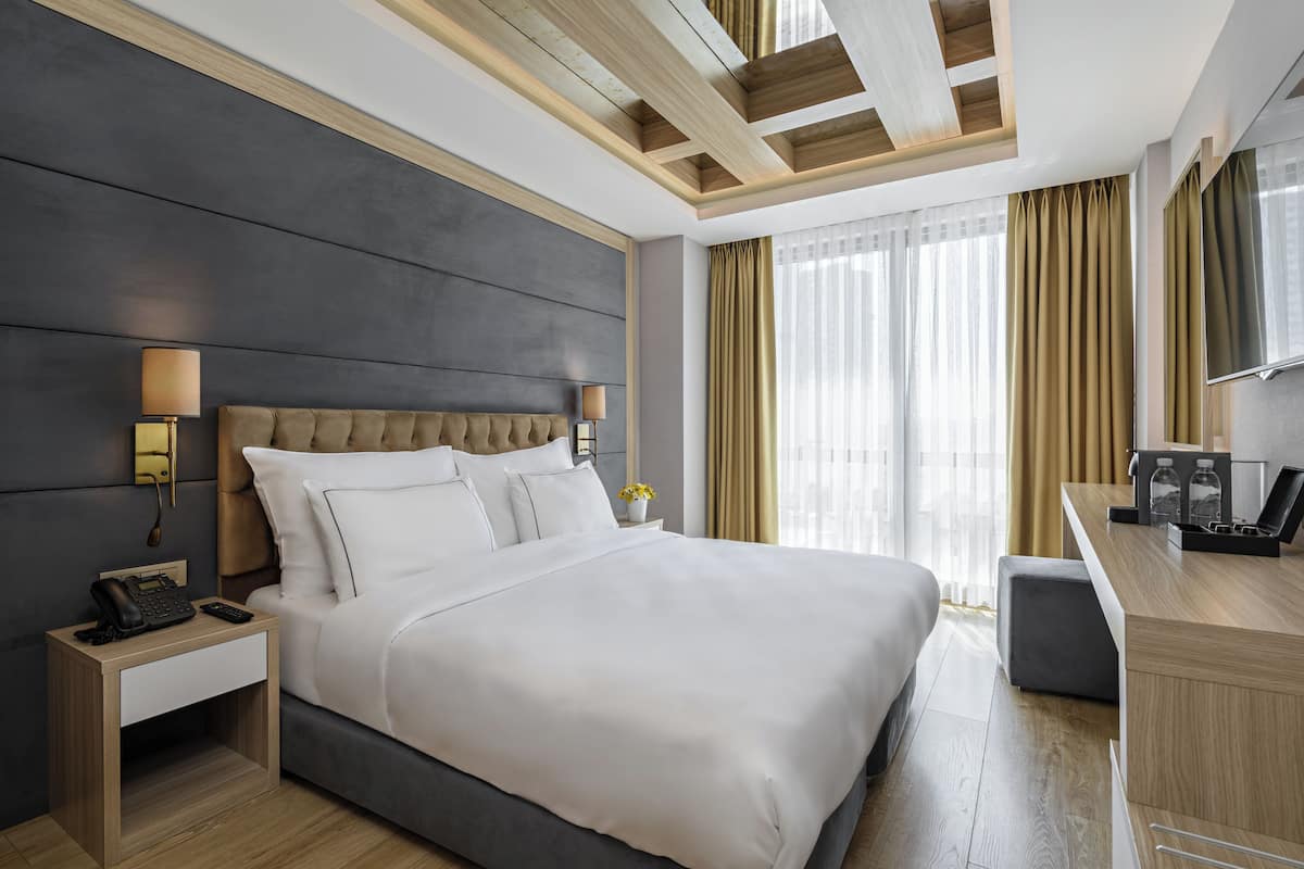 Habitación de hotel moderna con una cama grande, lámparas de noche, un escritorio con teléfono y una ventana con cortinas.