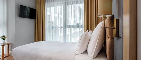 A modern hotel room with a large bed, a TV mounted on the wall, a bedside table with a remote control, and a window with sheer curtains.