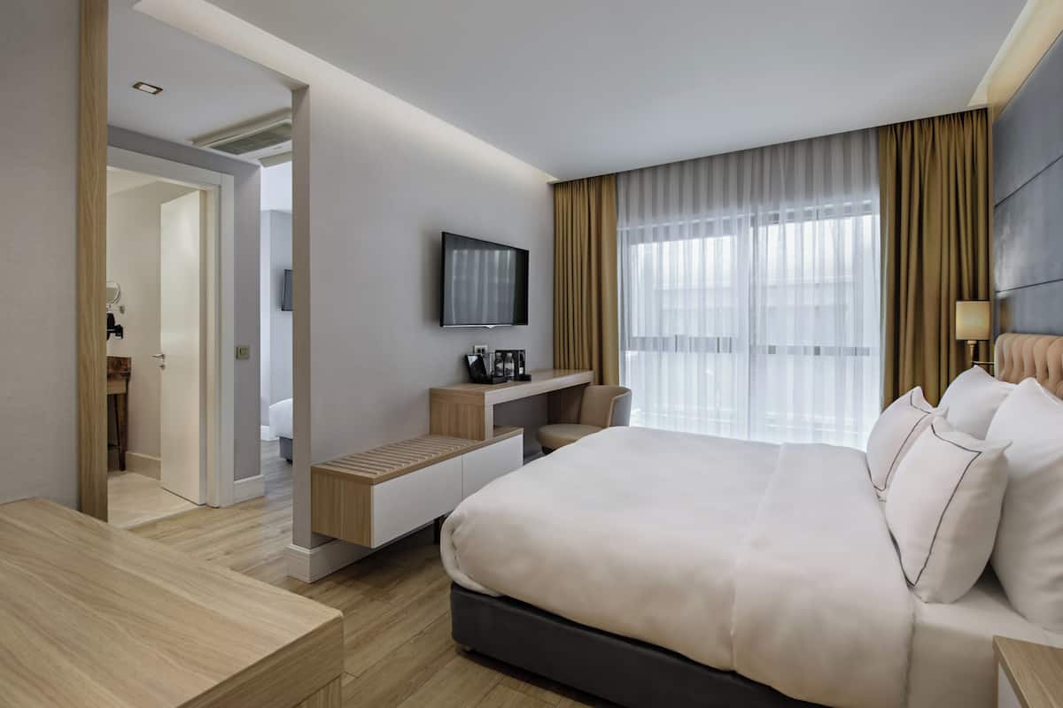 Una habitación de hotel moderna con una cama grande, un televisor montado en la pared, un escritorio con una silla y una ventana con cortinas traslúcidas.