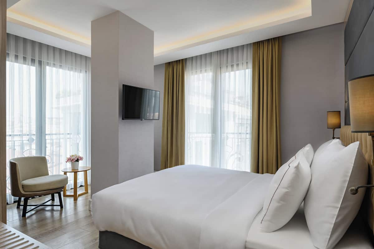 Una habitación de hotel moderna con una cama grande, un televisor de pantalla plana montado en la pared, una silla, una mesita con un jarrón de flores y ventanas amplias con cortinas.