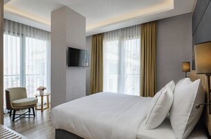 Una habitación de hotel moderna con una cama grande, un televisor de pantalla plana montado en la pared, una silla, una mesita con un jarrón de flores y ventanas amplias con cortinas.