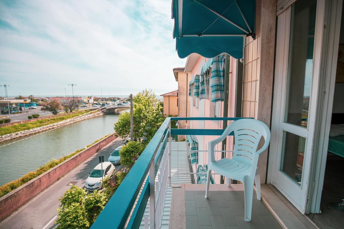 A balcony with a view of a canal, a white plastic chair, and a building with striped awnings.
