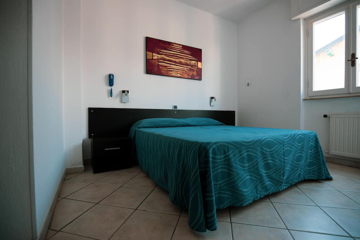 A bedroom with a bed, a nightstand, a wall-mounted phone, and a window.