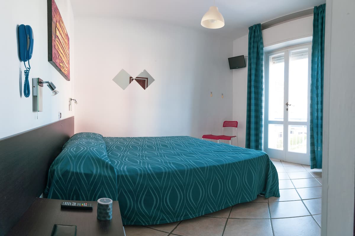 A hotel room with a bed, a desk with a remote control, a phone, a wall-mounted TV, and a window with curtains.
