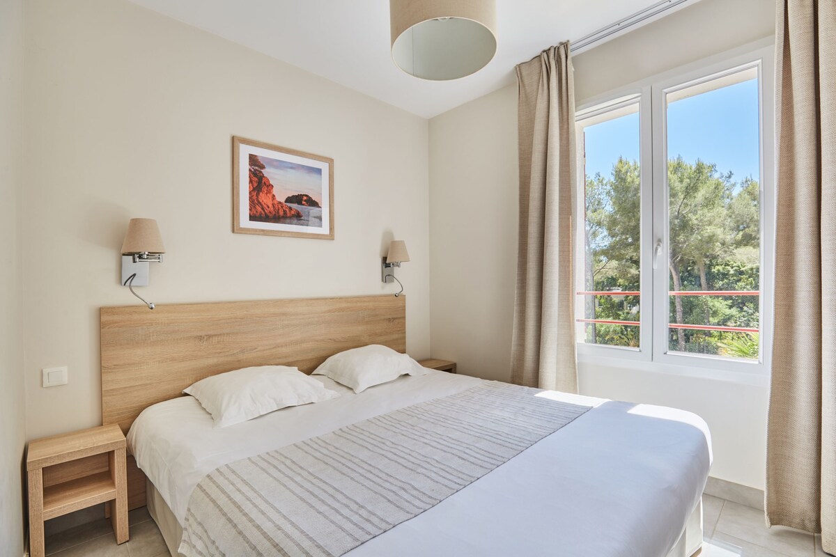 A bedroom with a wooden headboard, two bedside tables with lamps, a large window with curtains, and a framed picture on the wall.