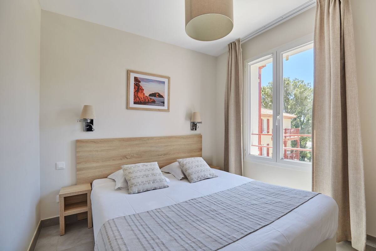 Ein Schlafzimmer mit hölzernem Kopfteil, zwei Nachttischen, einem Bett mit gemusterten Bettbezügen, einem gerahmten Bild an der Wand und einem Fenster mit Vorhängen.