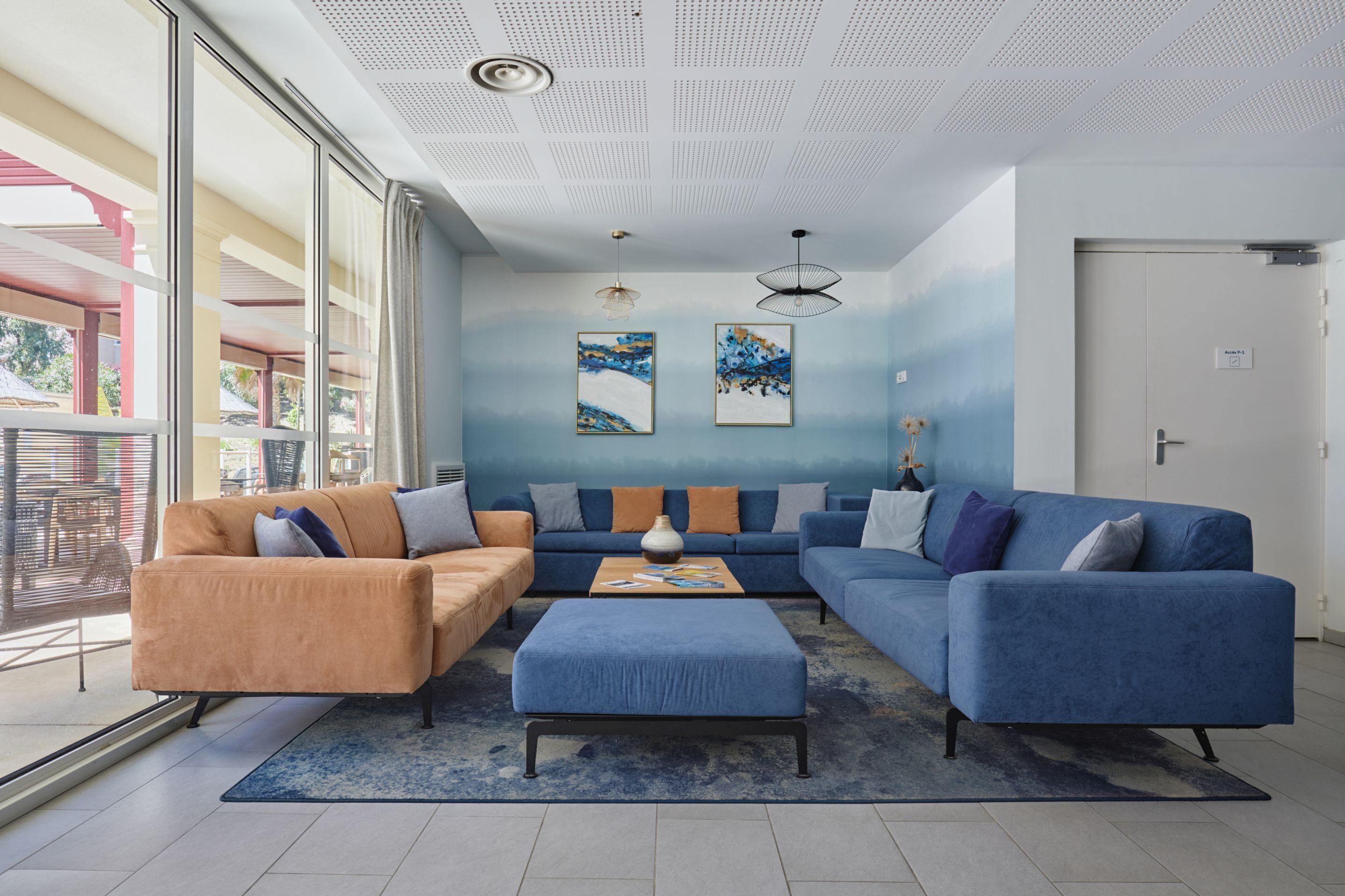 A modern living room with a blue sofa, an orange sofa, and a blue ottoman. There are two paintings on the wall, a ceiling fan, and a door with a sign.