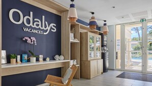 Un área de recepción con un letrero que dice "Odaly Vacances", estanterías de madera con objetos decorativos, sillas de mimbre e iluminación colgante.