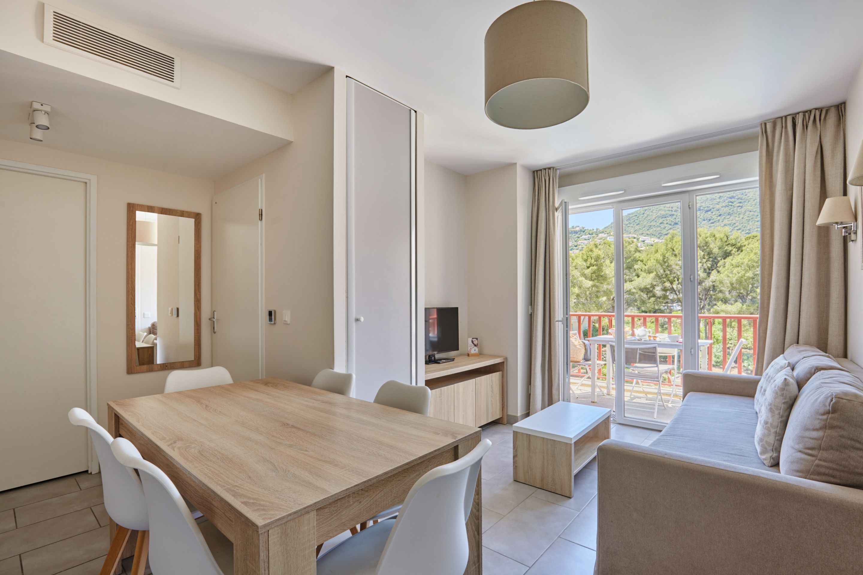 Une chambre d’hôtel moderne avec une table à manger en bois, des chaises blanches, un canapé, une télévision et un balcon offrant une vue sur la verdure.