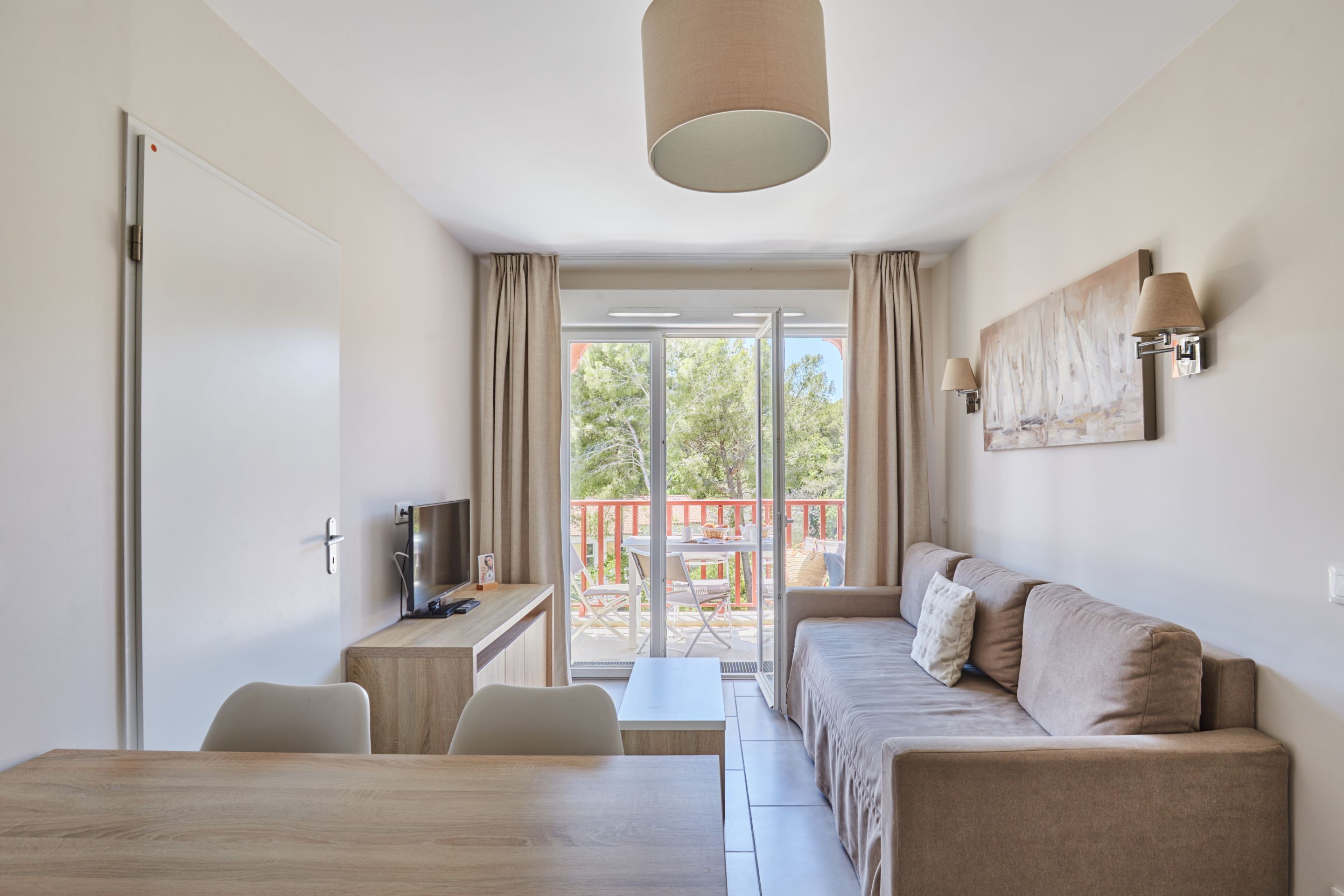 A modern room with a bed, a desk, a TV, and a balcony with a view of trees and a red railing.