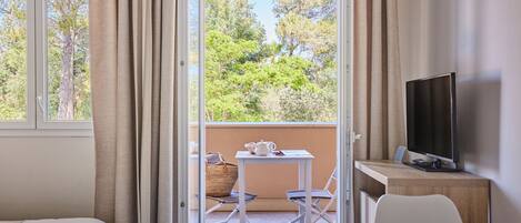 Ein Zimmer mit Balkon, einem Bett, einem Schreibtisch, einem Fernseher und Blick auf Bäume.
