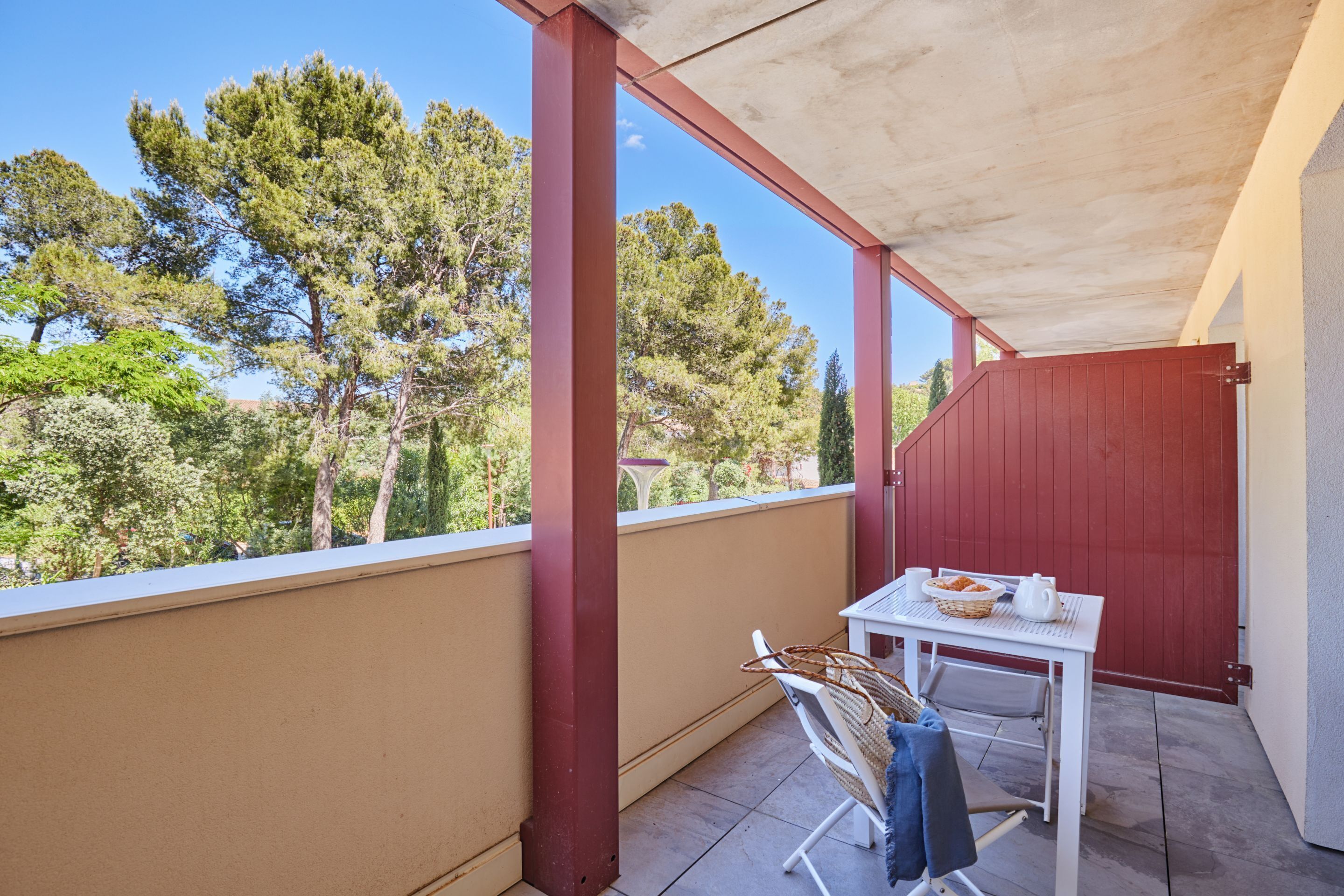 A covered outdoor seating area with a table and chairs, overlooking a garden with trees.