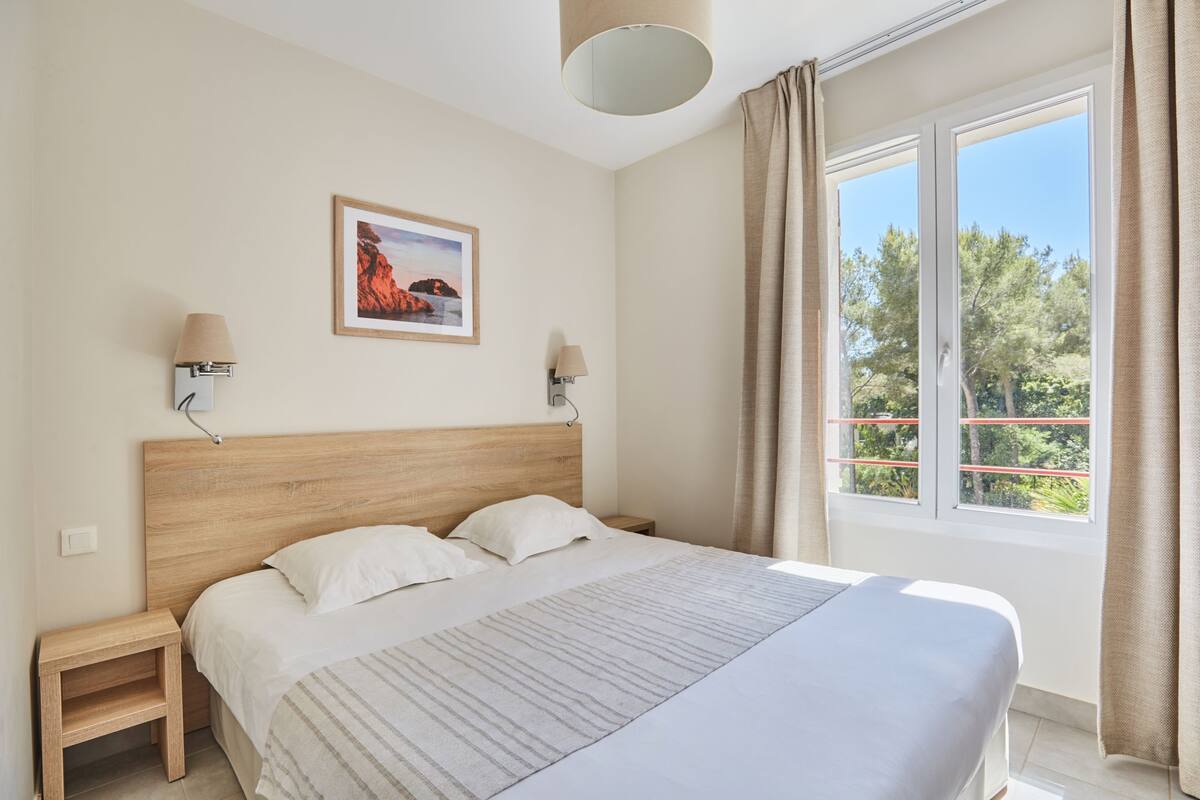 Ein Schlafzimmer mit einem Holzkopfteil, zwei Nachttischen mit Lampen, einem großen Fenster mit Vorhängen und einem gerahmten Bild an der Wand.