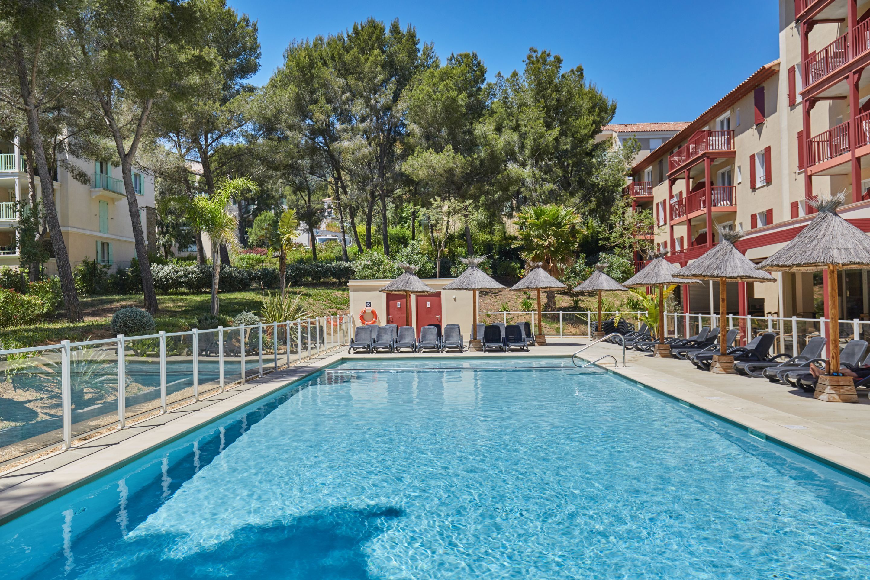 A swimming pool with lounge chairs and umbrellas, surrounded by trees and a building with balconies.