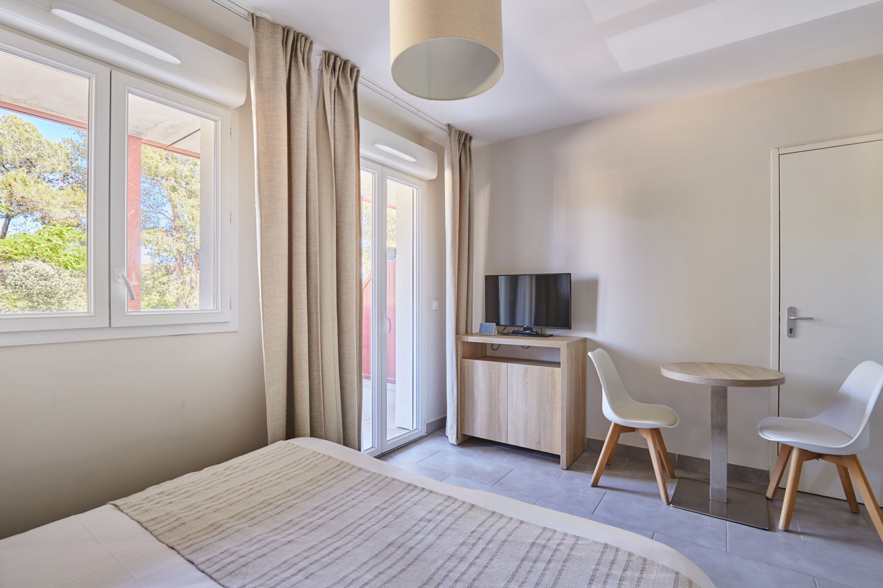 A hotel room with a bed, a desk with a TV, two chairs, and large windows with curtains.