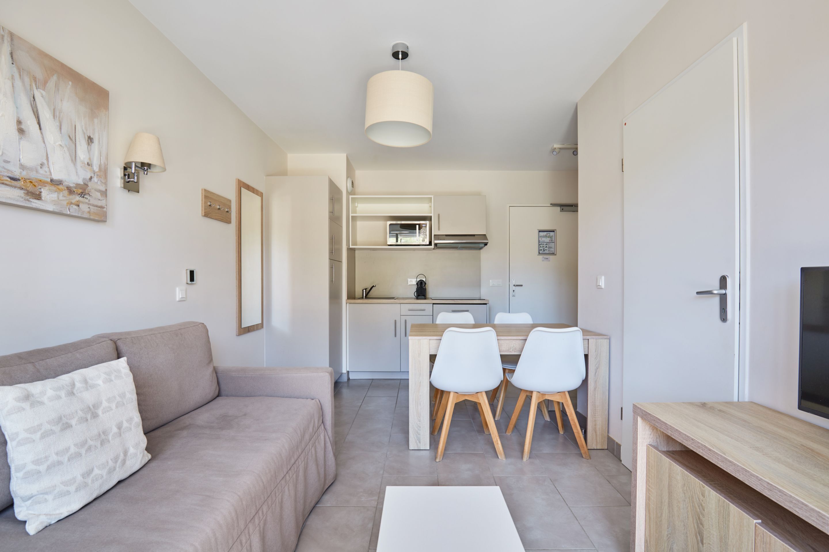 A modern living room with a sofa, a dining table with chairs, a kitchen area with cabinets and appliances, and a TV on a wooden stand.