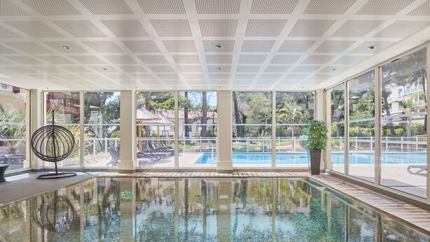 Una piscina interior con paredes de vidrio transparente, un techo con patrón de cuadrícula y vistas a una zona de piscina exterior.