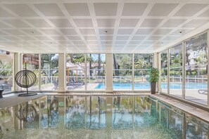 Une piscine intérieure avec des parois en verre transparent, un plafond à motifs géométriques et une vue sur l’espace extérieur dédié à la piscine.