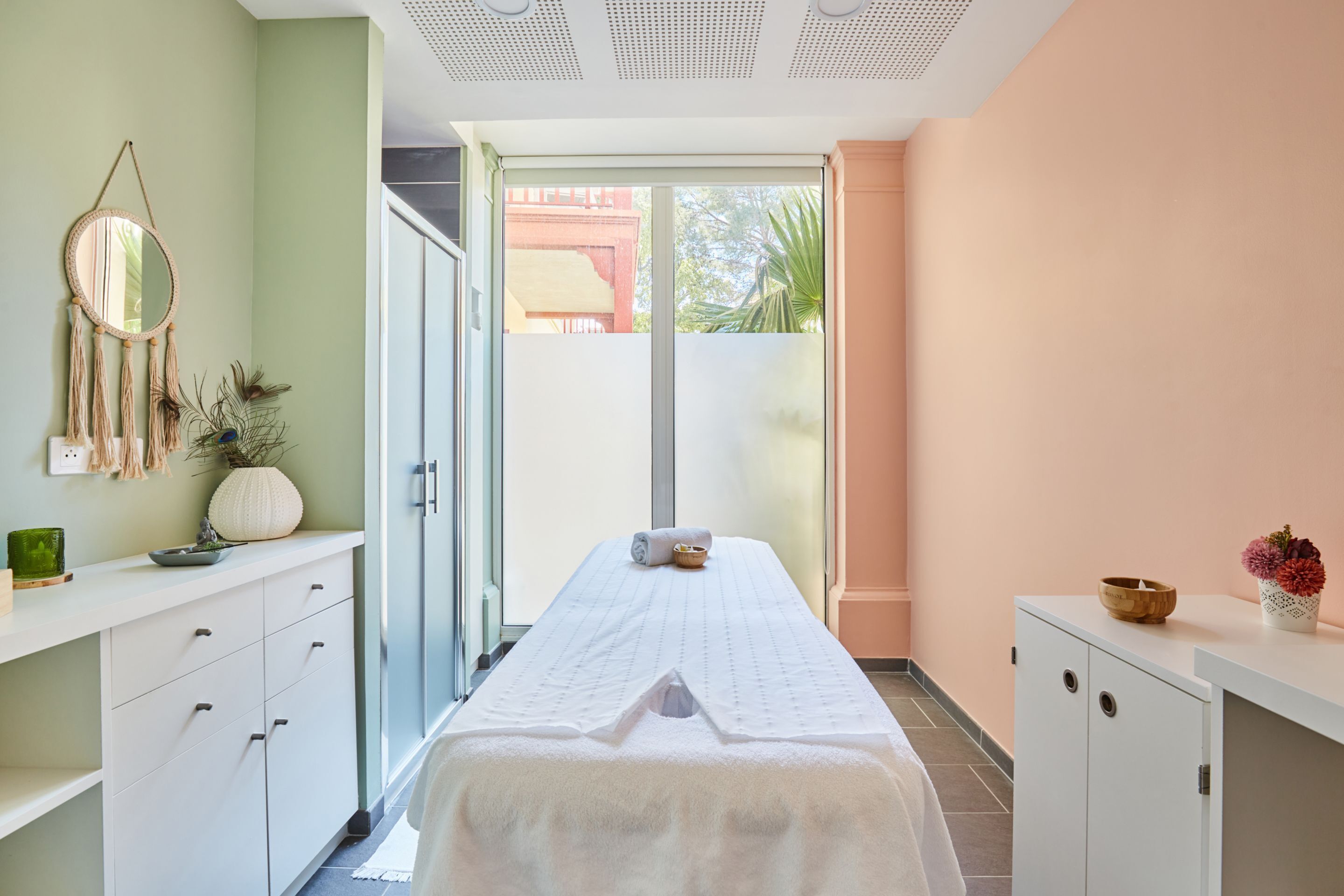 A spa room with a massage table, a vanity with a mirror, and a window with a view of greenery.