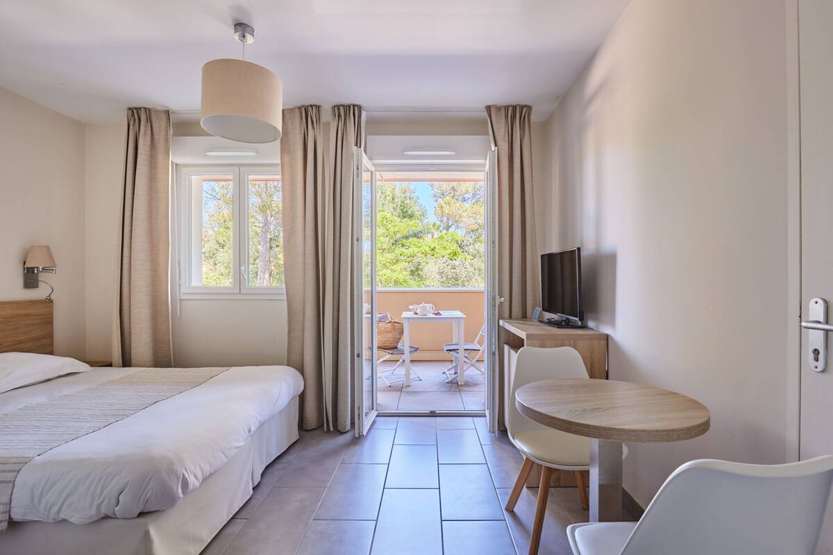 Ein Hotelzimmer mit Bett, Fernseher, einem kleinen Tisch und einem Balkon mit Blick auf Bäume.