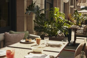An outdoor dining area with a table set for two, featuring a bowl of cereal, a glass of water, and a plate with a slice of cake. There are potted plants and a wicker basket in the background. - The Hoxton, Downtown LA (Los Angeles)