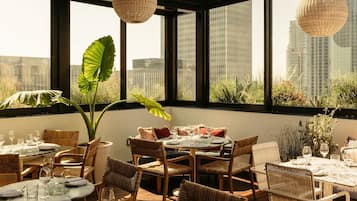 Un espace repas avec des tables rondes et des chaises en osier, de grandes fenêtres offrant une vue sur la ville et des lampes suspendues en osier.