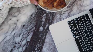 Una persona sostiene un plato con un croissant, dos recipientes pequeños de mermelada y una taza roja con un café con leche sobre una mesa de mármol.