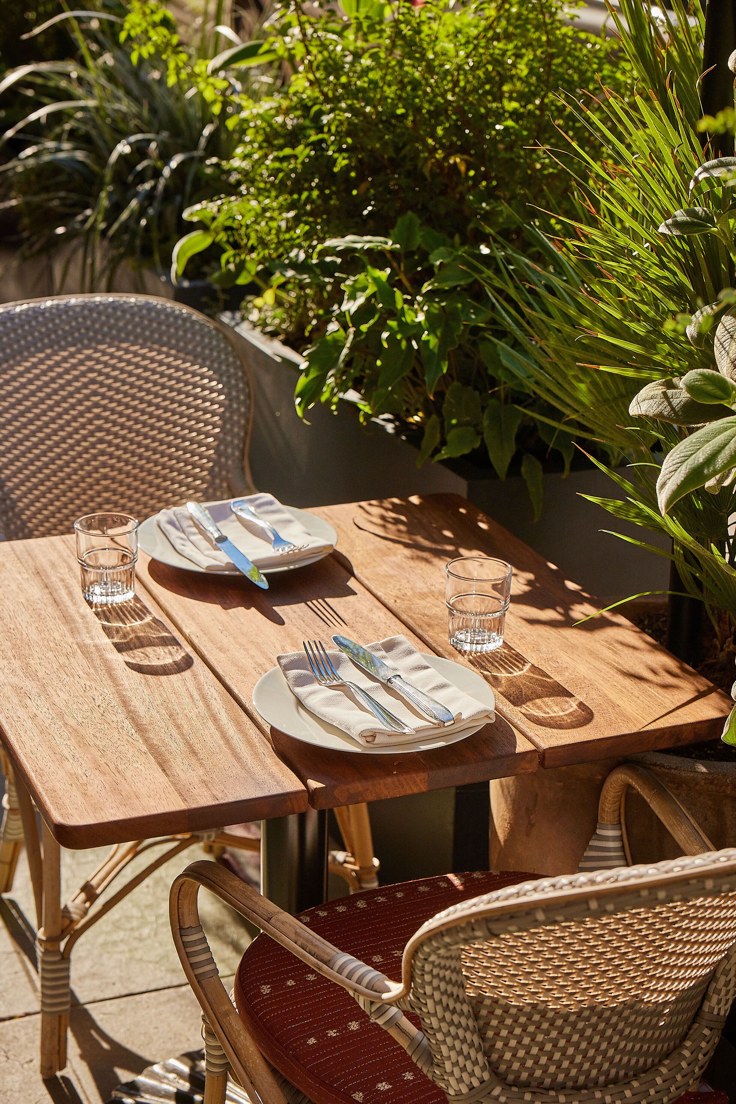 Een houten tuintafel gedekt met borden, bestek en glazen, omringd door rieten stoelen en weelderige begroeiing.