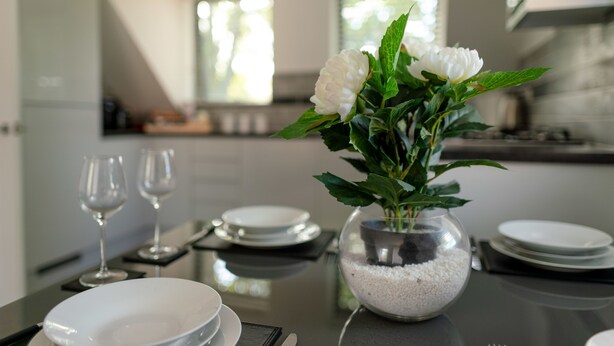Ein Esstisch ist gedeckt mit Tellern, Gläsern und einer Vase mit weißen Blumen.