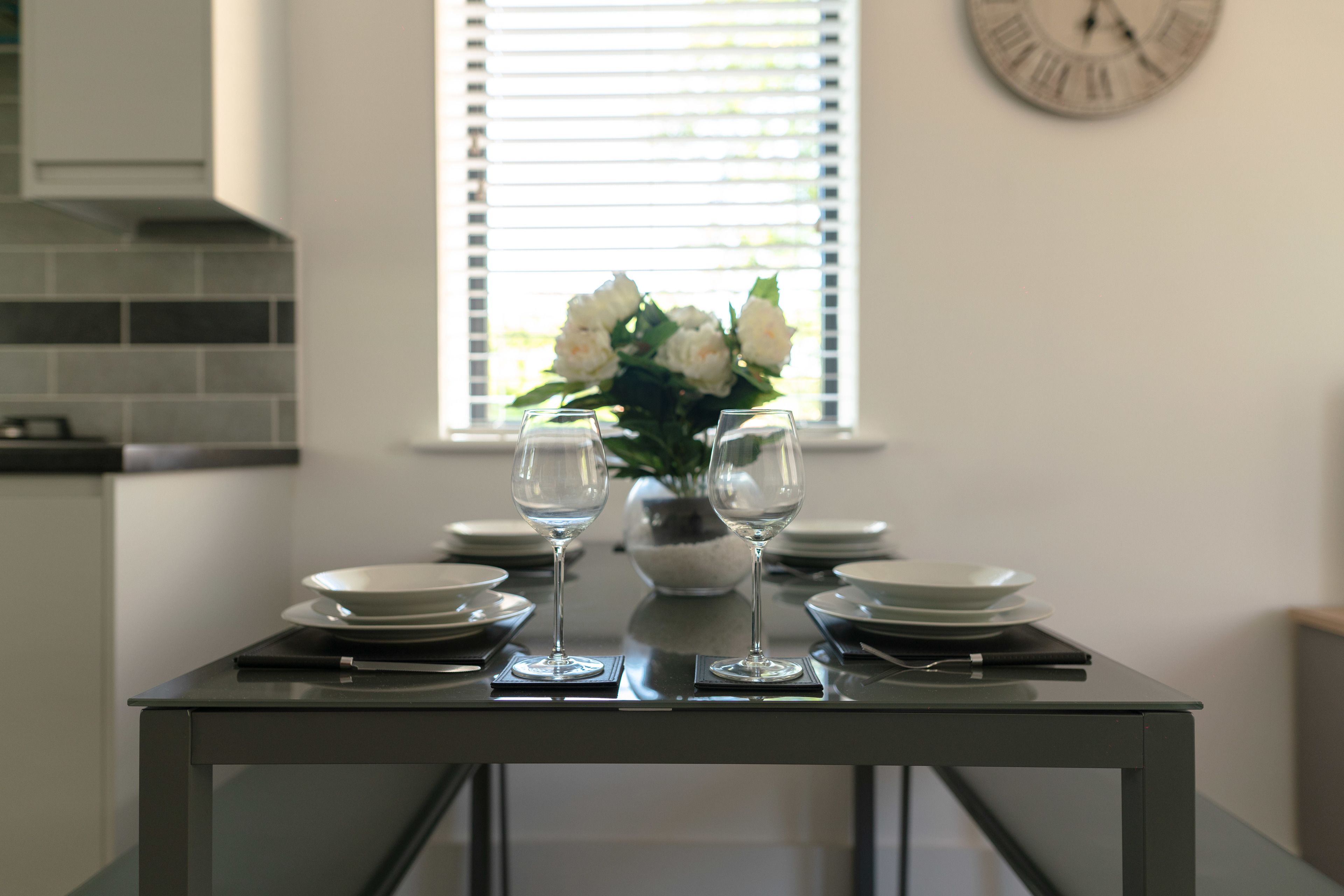 A dining table with two wine glasses, a vase with flowers, and plates set for two.