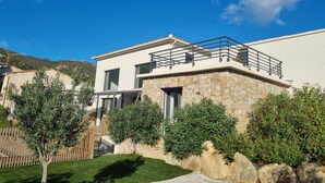 A modern two-story house with a stone facade, a balcony with a metal railing, and a well-maintained garden with various shrubs and a small tree. - Architect-designed villa, secure private pool, near center and beach  (Calvi)
