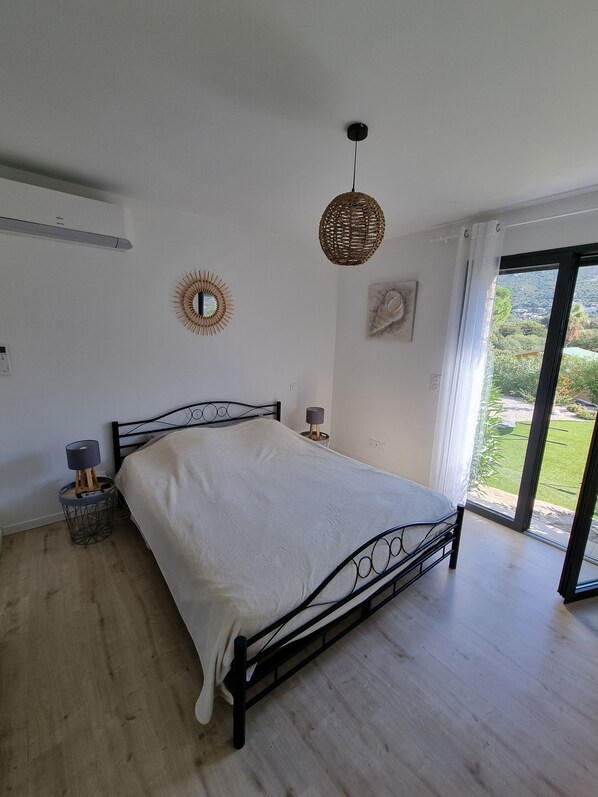 A bedroom with a bed, a bedside lamp, a wall-mounted mirror, and a view of a garden through a window. - Architect-designed villa, secure private pool, near center and beach (Calvi)