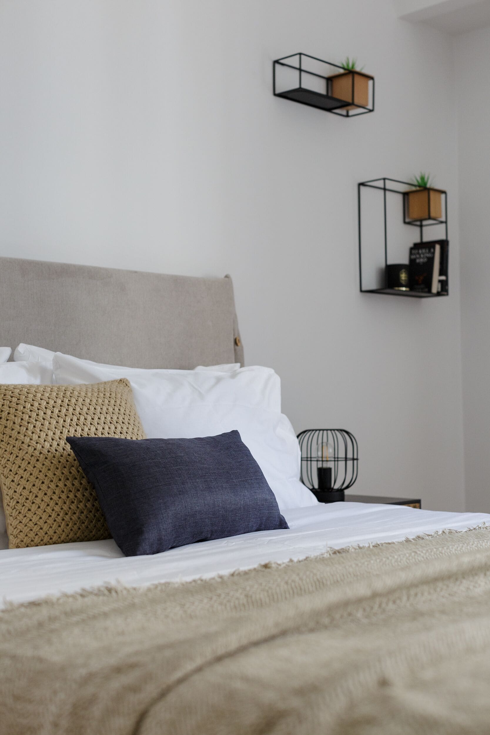 Un lit bien fait, recouvert d’une couverture beige, avec un oreiller bleu foncé et un plaid en tissu tissé. Deux étagères murales sont fixées au mur, ornées de plantes et d’un petit objet décoratif.