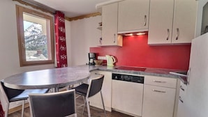 A compact kitchen with a round dining table, two chairs, a dishwasher, and a red accent wall. - 150m from ski slopes, floor -2, view mountain, balcony, 37m², Méribel-Mottaret (Méribel - Les Allues)