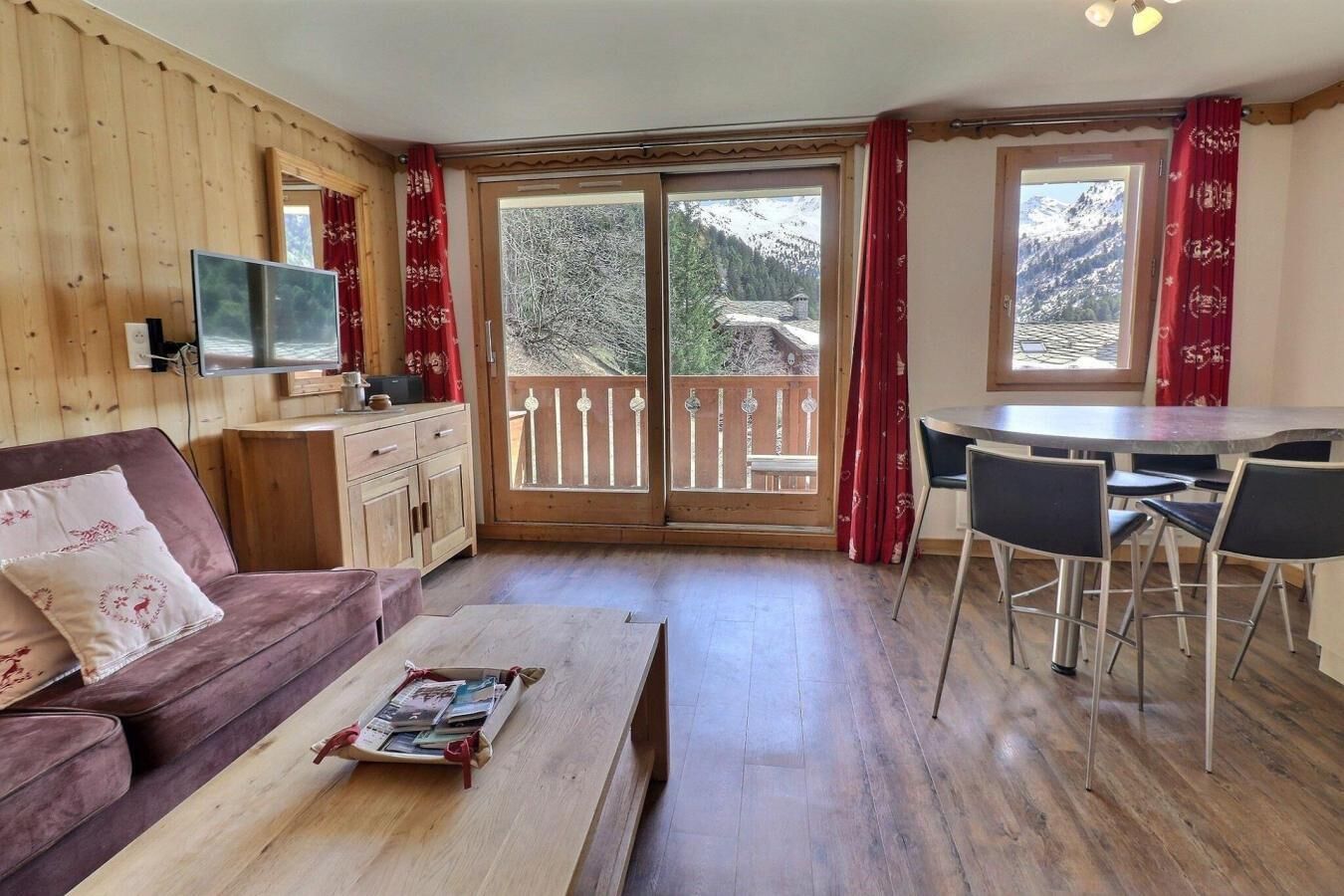Ein gemütliches Zimmer mit Holzvertäfelung, einer Couch, einem Couchtisch, einem Fernseher, einem Balkon mit Holzgeländer und einem Essbereich mit Stühlen.