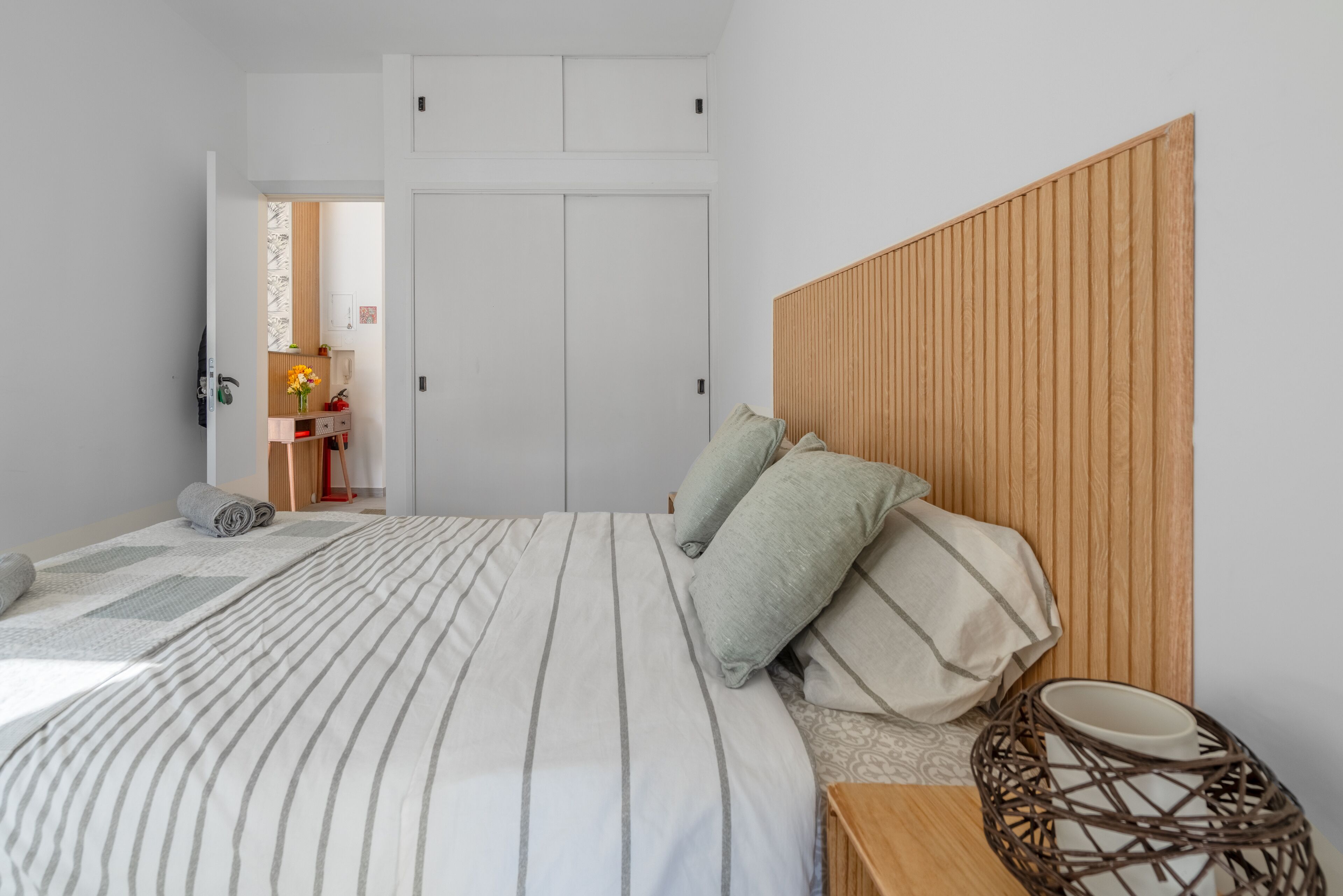A bedroom with a bed, pillows, a bedside table, a wooden headboard, a wardrobe, and a small table with a vase.