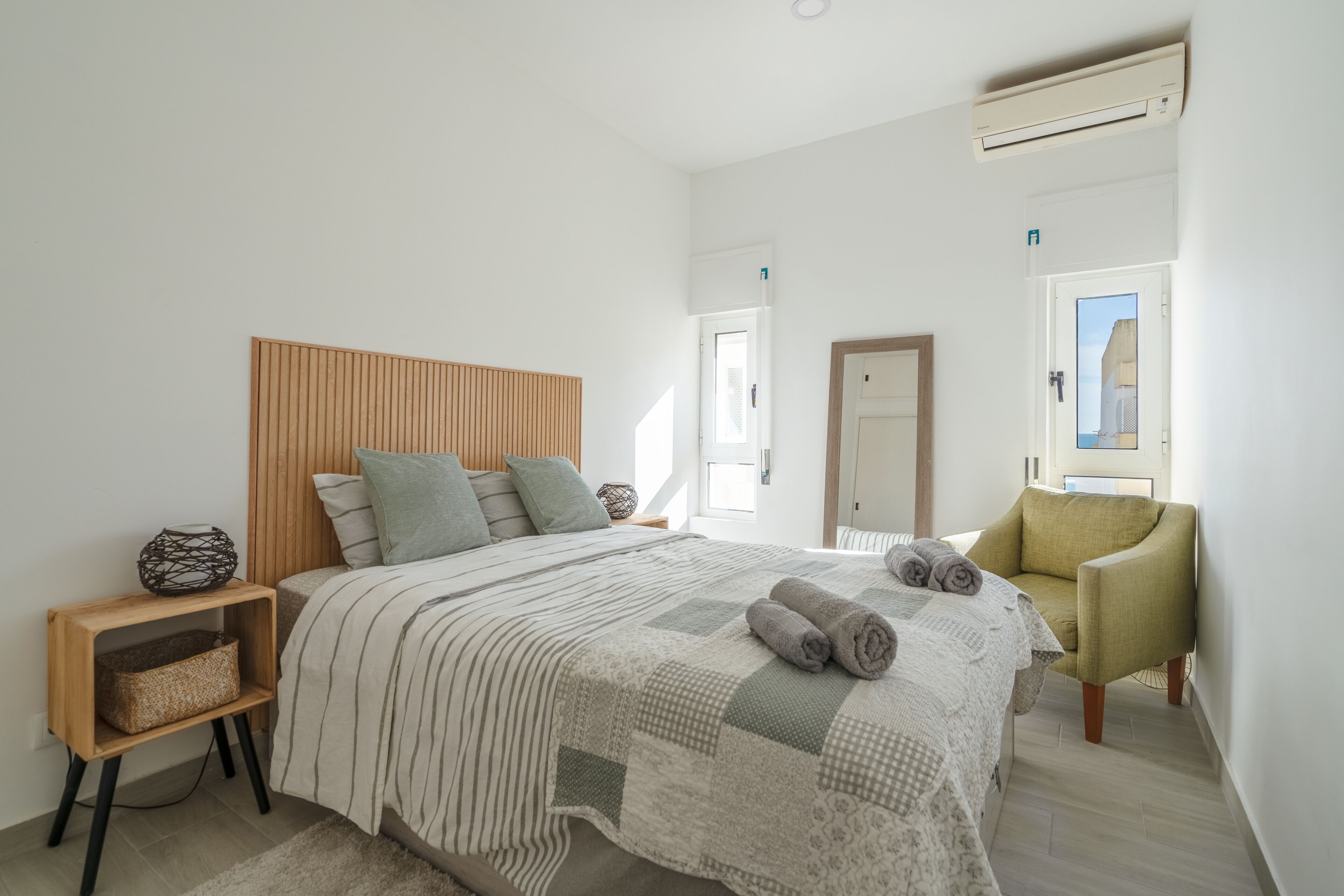 A modern bedroom with a wooden headboard, a bed with striped bedding, a green armchair, and a bedside table with a woven basket.