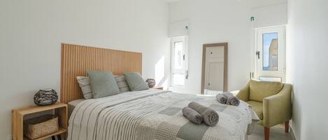 A modern bedroom with a wooden headboard, a bed with striped bedding, a green armchair, and a bedside table with a woven basket.