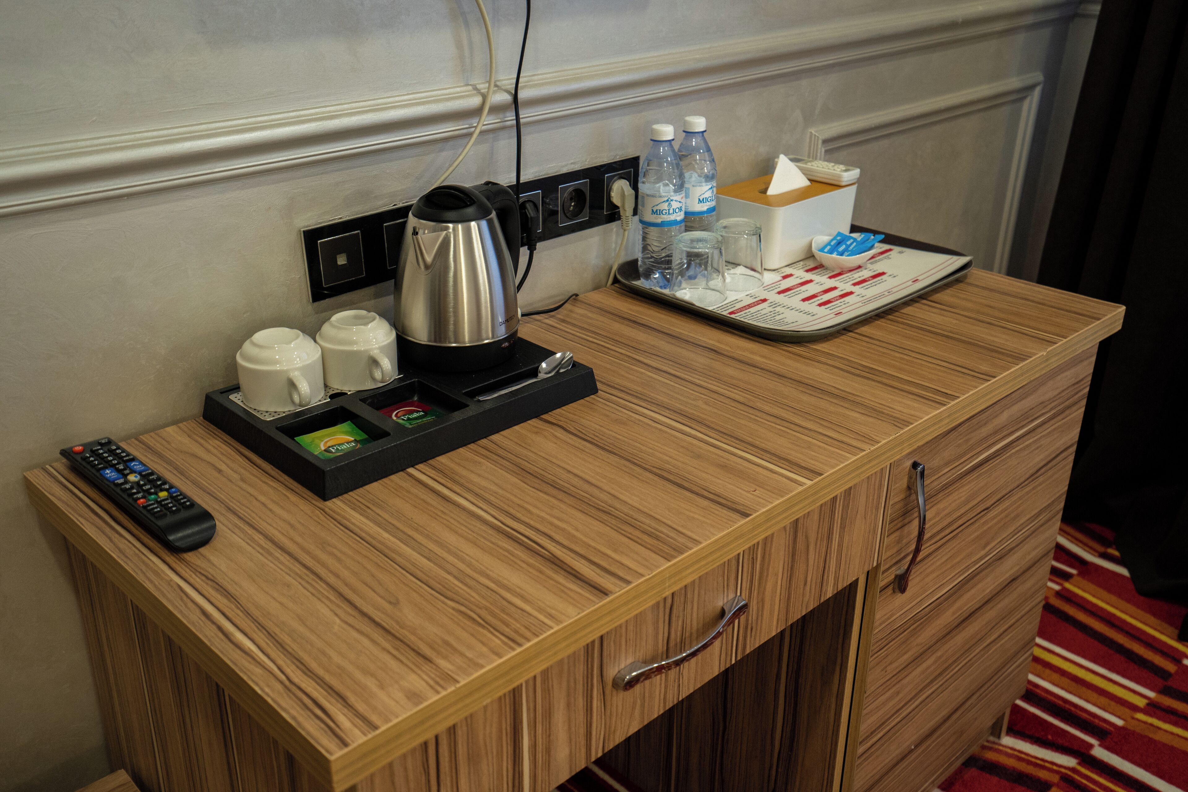 A wooden desk with a remote control, a kettle, and a tray with water bottles and a tissue box.