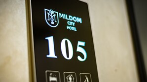 A digital display with the MILDOM CITY HOTEL logo and room number 105. - Mildom City Hotel (Almaty)