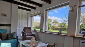 Una sala de estar con sofá, sillón y una mesa. Se ve un ventanal con vistas a zonas verdes.