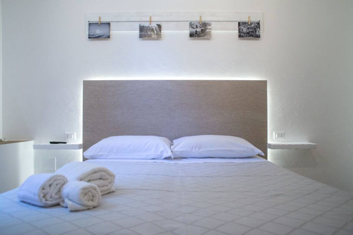 Une chambre d’hôtel avec un grand lit, du linge de lit blanc et une tête de lit. Il y a trois petits tableaux accrochés au mur au-dessus du lit.