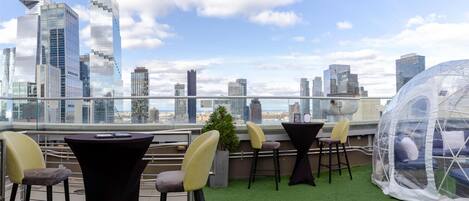 A rooftop terrace with a city skyline view, green artificial turf, and outdoor seating with chairs and tables.