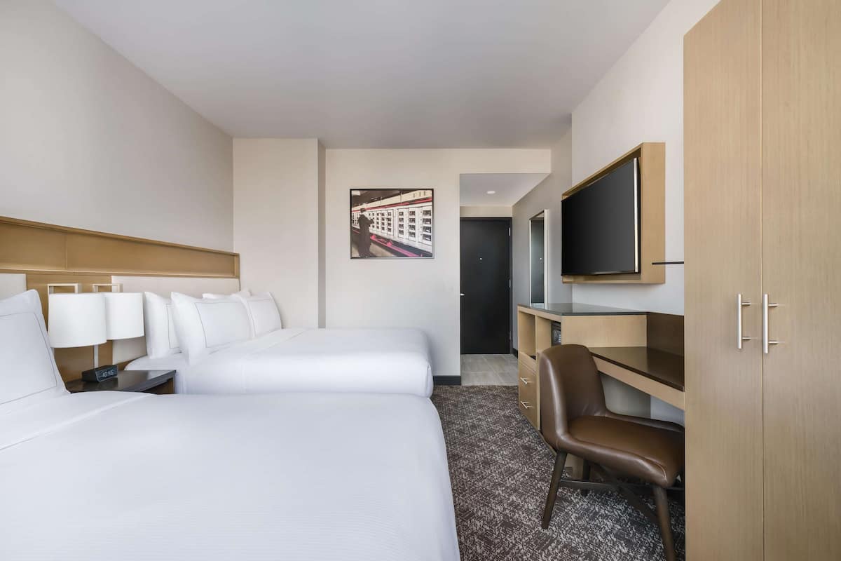 Ein Hotelzimmer mit zwei Betten, einem Schreibtisch, einem Sessel, einem Fernseher und einem Bild an der Wand.
